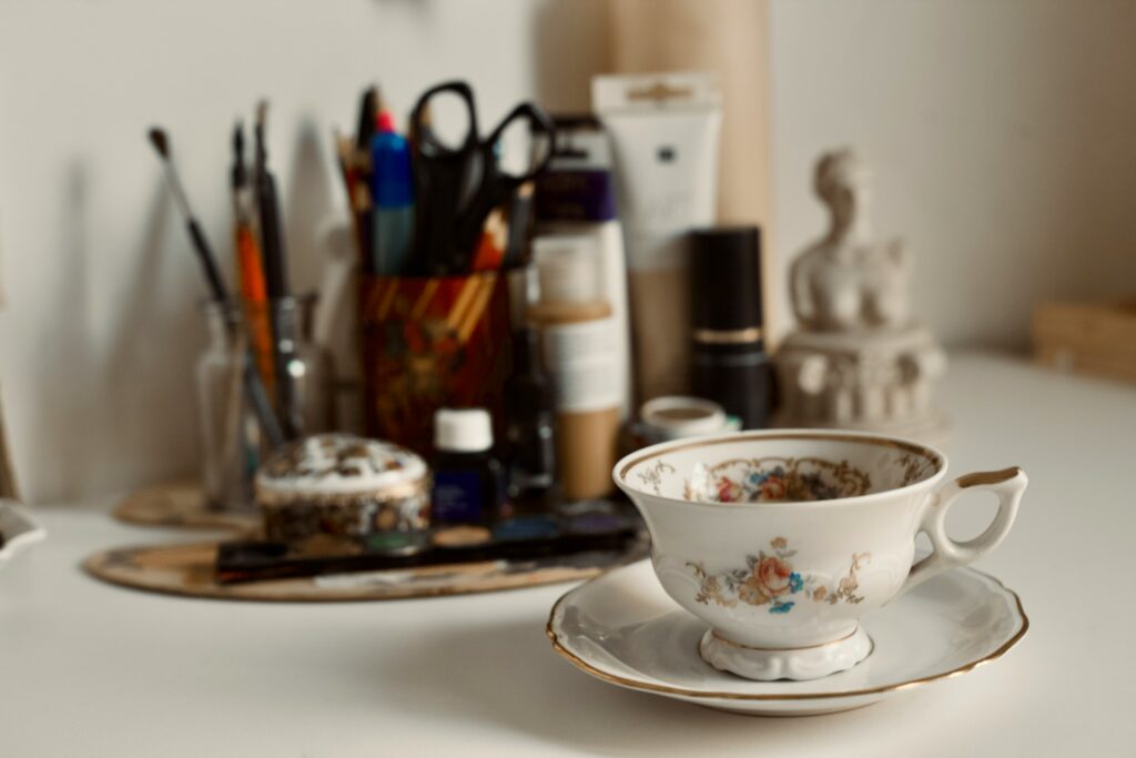 Close up of an antique floral teacup and saucer resting on a white desk with art supplies, a paint palette, and a small sculpture blurred in the background, representing the careful curation process of Porcelain Vignette.