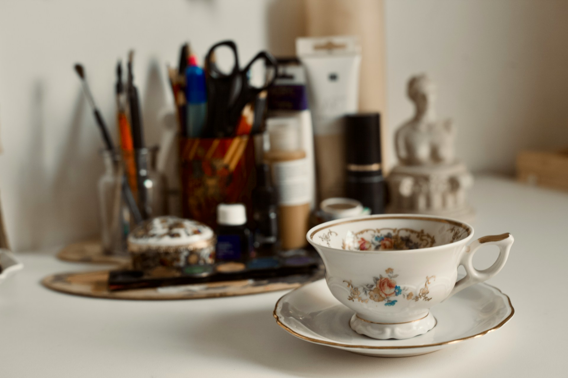 Close up of an antique floral teacup and saucer resting on a white desk with art supplies, a paint palette, and a small sculpture blurred in the background, representing the careful curation process of Porcelain Vignette.