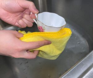 Close-up of hands gently polishing a gold-rimmed floral teacup using a soft yellow microfiber cloth to remove tarnish and restore shine without abrasives