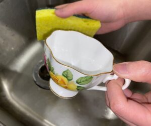 A person hand-washing a delicate vintage teacup with a soft yellow sponge in a kitchen sink, demonstrating the proper technique for preserving overglaze gold accents